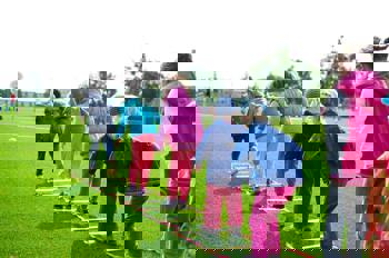 Children in colourful jackets play outdoors, balancing on a ladder laid flat on green grass, showcasing teamwork and fun on a bright day.