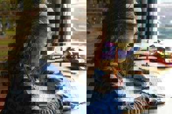 A young child with long hair is focused on doing homework at a wooden table. They're holding a pencil, colouring in a worksheet. 