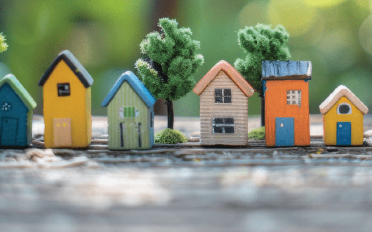Miniature colourful houses with varied designs are lined up on a wooden surface, surrounded by small green trees.