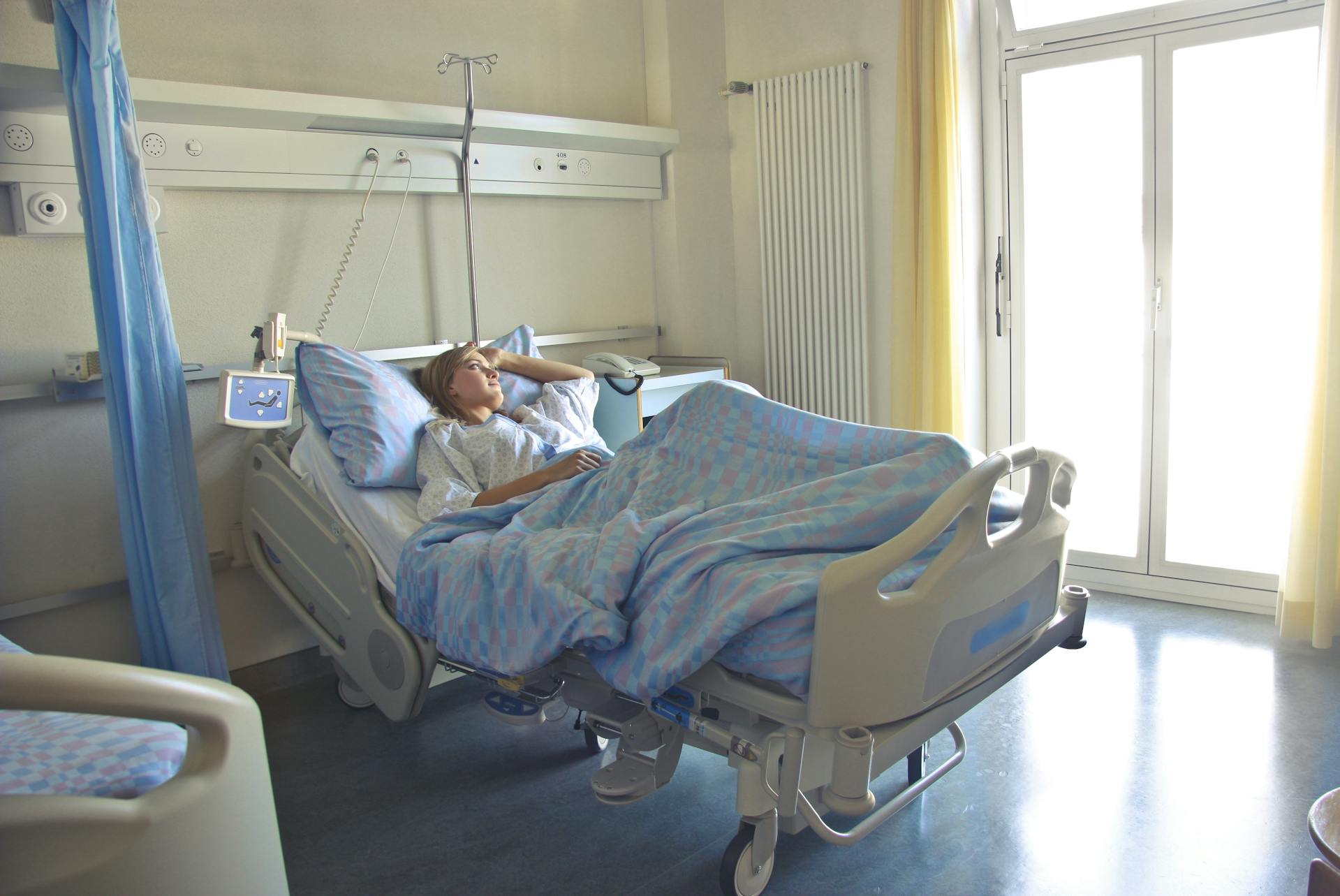 A person rests in a hospital bed, looking contemplative. Sunlight filters through large windows, creating a calm, serene atmosphere in the room.