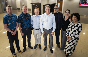 A group of seven people posing together in a modern building hallway.