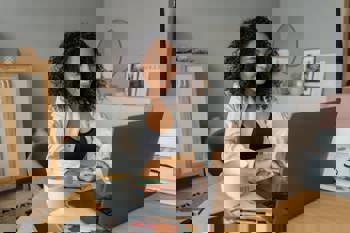 Smiling woman with curly hair works on a laptop at home. She's seated at a wooden table with a coffee mug, papers, and stationery, creating a cosy, focused ambiance.