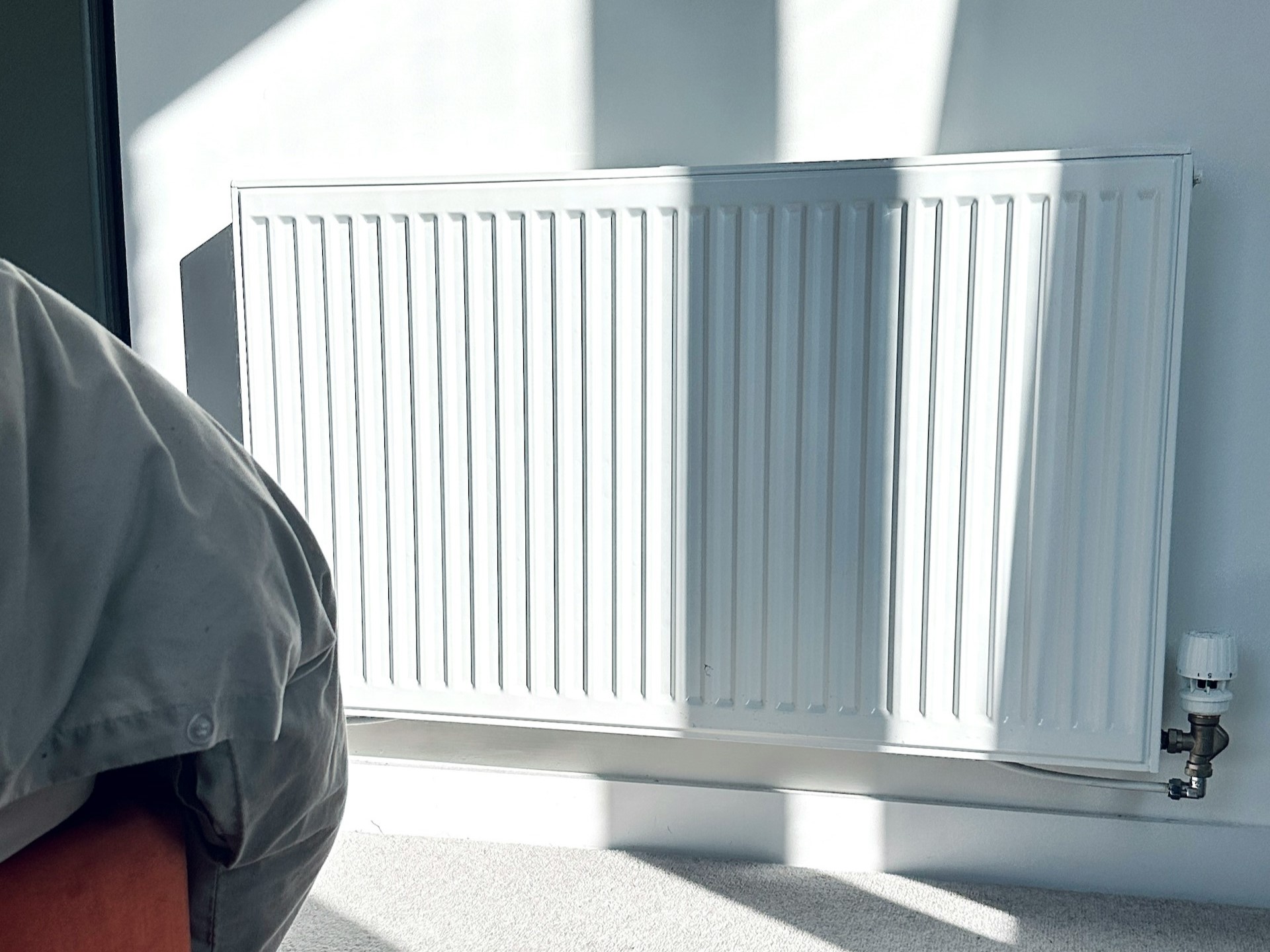 A white radiator casts shadows on a wall, with a corner of a beige fabric sofa partially visible on the left. Sunlight streams in.