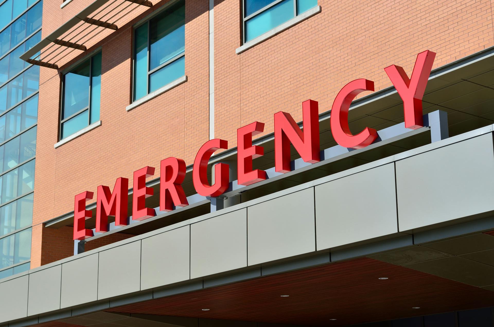 Red "EMERGENCY" sign on a hospital building facade with large windows and brickwork