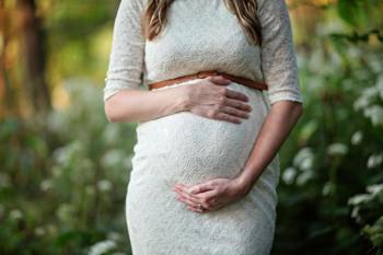 Pregnant woman in lace dress gently holds her belly with both hands, standing in a serene, green woodland setting.
