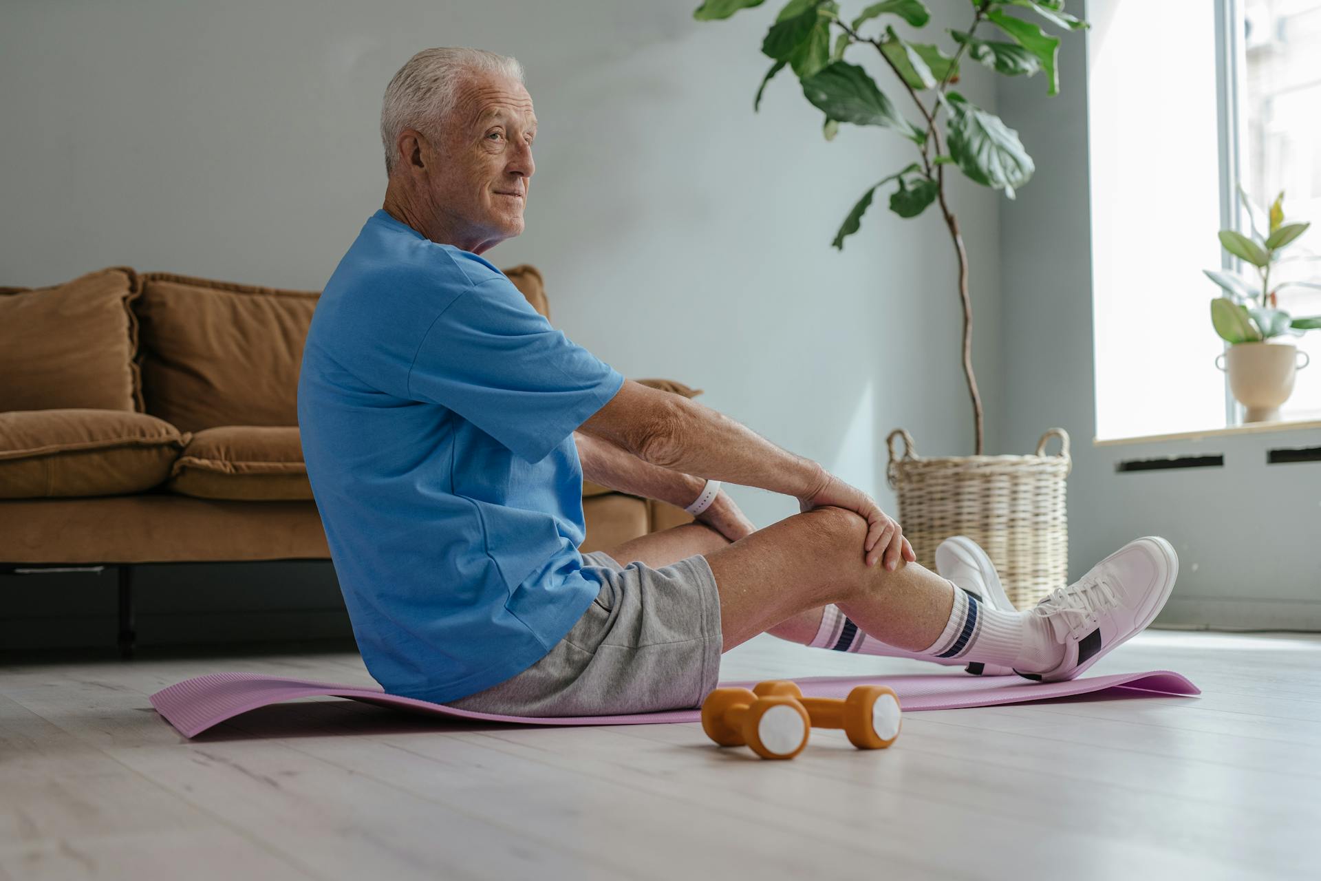Elderly man in a blue shirt and shorts sits on a yoga mat in a living room, stretching and smiling. Nearby, orange dumbbells and a large potted plant.