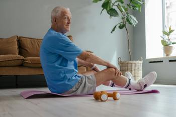 Elderly man in a blue shirt and shorts sits on a yoga mat in a living room, stretching and smiling. Nearby, orange dumbbells and a large potted plant.