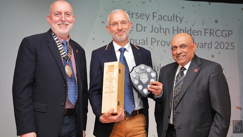 Three men in suits stand together, smiling. The man in the centre holds a trophy and shield. A screen in the background displays an academic award ceremony.