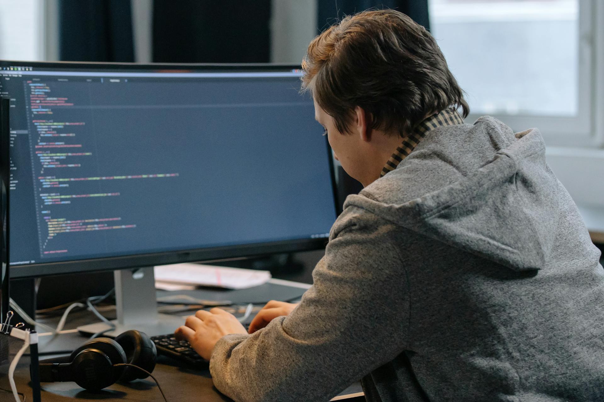 A person in a gray hoodie is focused on typing code on a large monitor. Headphones are placed nearby.