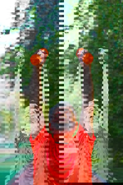 Elderly man exercising outdoors, lifting orange dumbbells with arms raised. He wears a bright red shirt, surrounded by greenery.