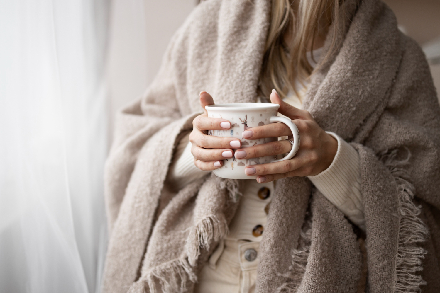 A woman wrapped in a cozy beige blanket holds a patterned mug with both hands. 